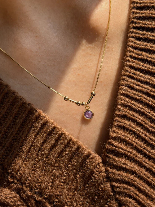 Close-up detail of a delicate 22K gold Morse code initial necklace with a 6mm purple birthstone charm, worn in warm natural sunlight[cite: 12].