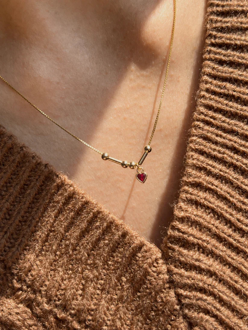Minimalist his and hers Morse code initial necklace crafted in 22K gold, featuring a tiny red 3mm heart stone worn beautifully in natural sunlight[cite: 10].