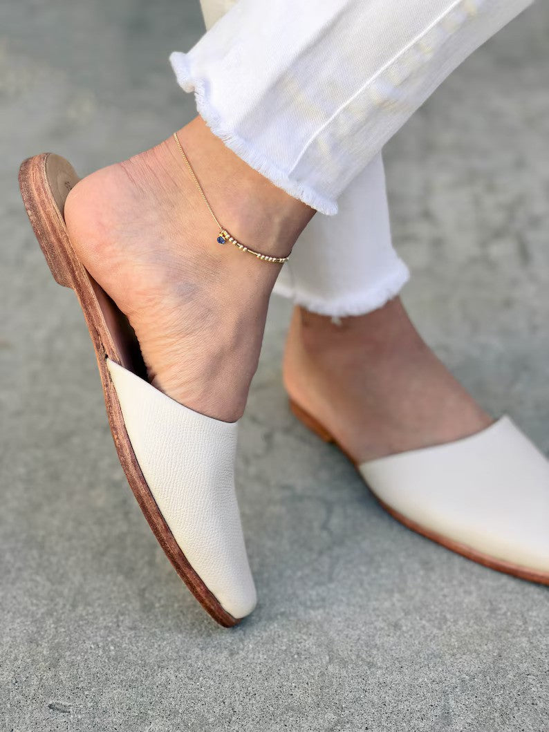 A close-up of a personalized gold Morse code name anklet adorned with a delicate birthstone charm, worn elegantly on a woman's ankle with white mules[cite