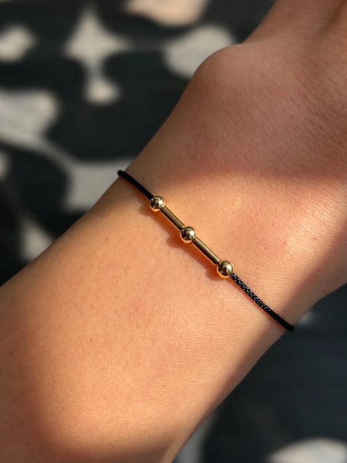 Detail shot of a personalized Morse code thread bracelet on a wrist, featuring 22K gold finish beads against a minimal black cord in natural sunlight.