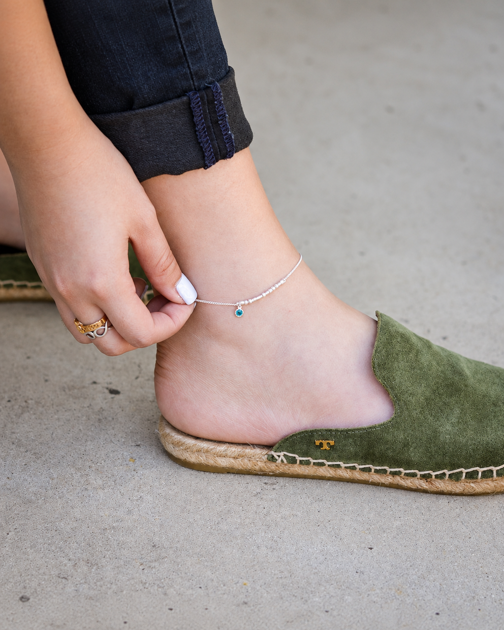 Close-up of a personalized silver Morse code anklet with custom initials and a tiny bezel-set birthstone charm, worn by a woman with casual footwear